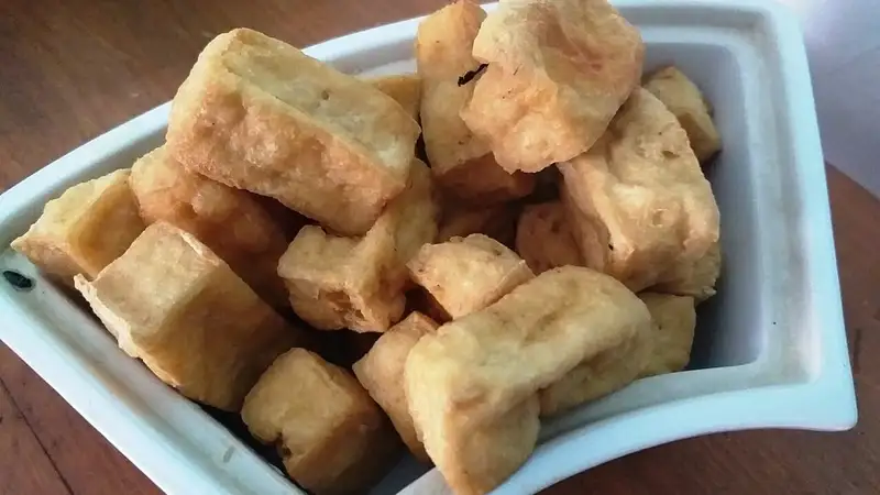 Potongan tahu goreng berwarna kuning keemasan bertumpuk di dalam piring saji putih.