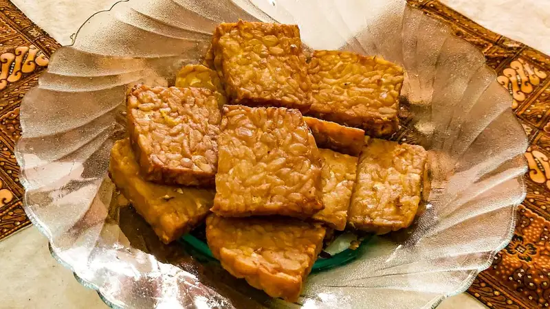 Potongan tempe goreng berwarna cokelat keemasan bertumpuk di atas piring kaca, dengan kain batik sebagai alas.