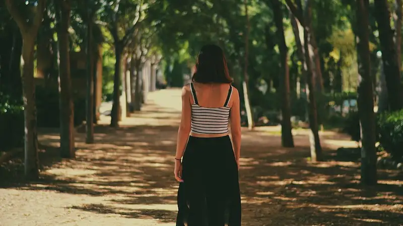 Seorang perempuan berjalan sendirian menyusuri jalan setapak di bawah deretan pohon rindang, dilihat dari belakang.