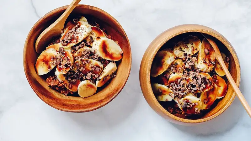 Dua mangkuk kayu berisi oatmeal dengan irisan pisang dan taburan kakao, dilihat dari atas di atas meja marmer putih.