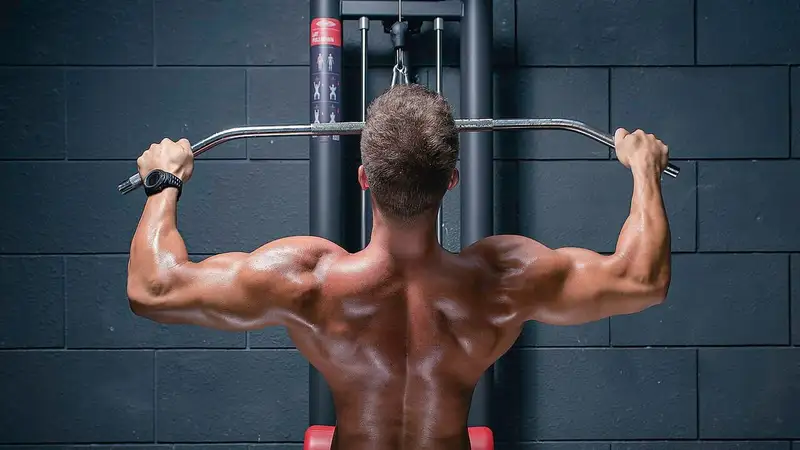 Seorang atlet terlihat dari belakang menarik palang mesin lat pulldown sehingga otot punggungnya tampak jelas