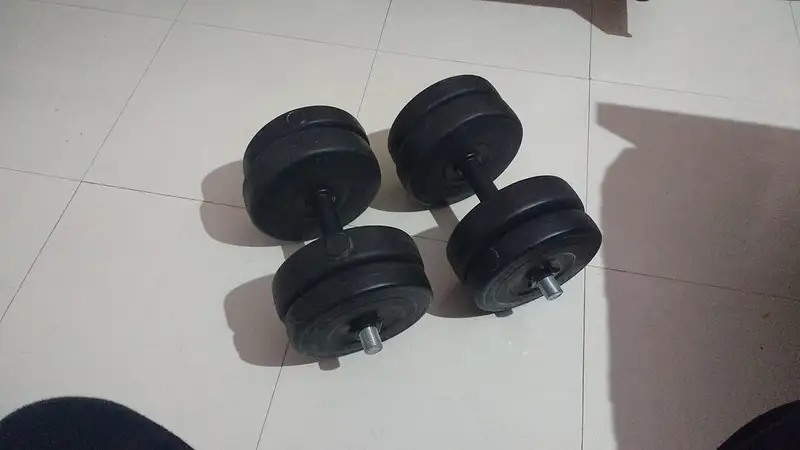 Sepasang dumbel hitam dengan piringan beban yang bisa dilepas pasang tergeletak di lantai keramik putih.