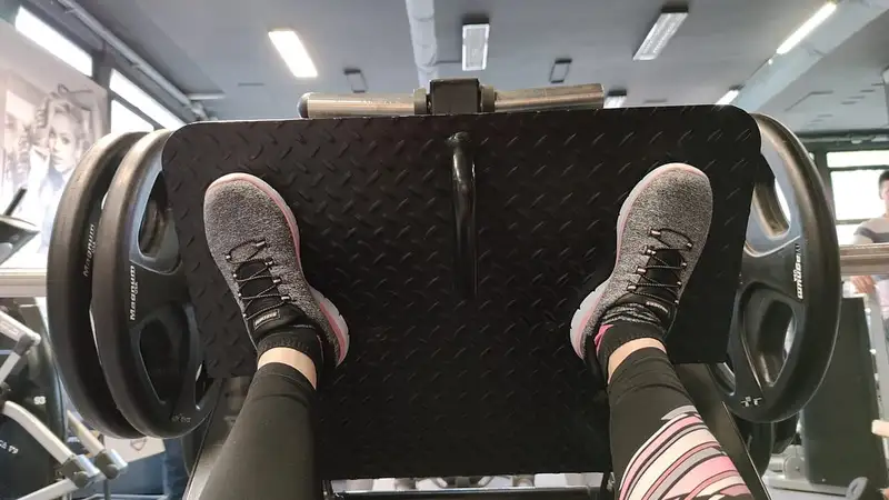 Sudut pandang orang pertama saat mendorong pijakan mesin leg press berisi lempeng beban dengan kedua kaki bersepatu olahraga.
