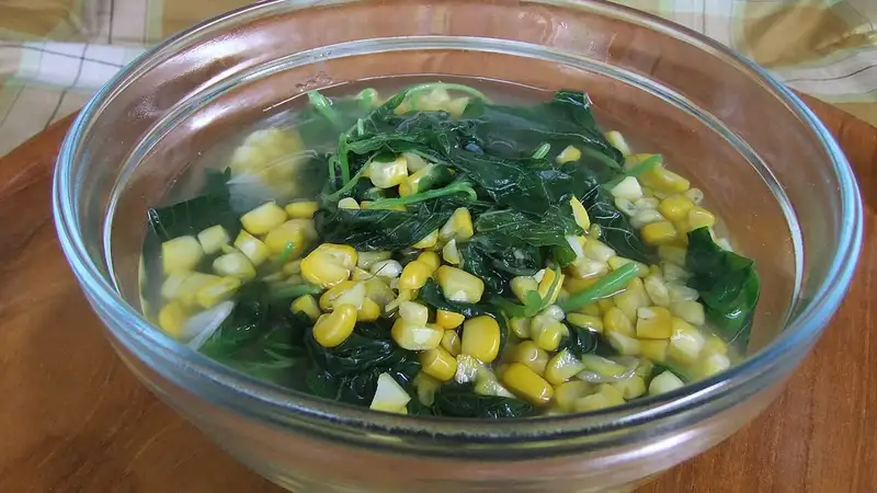 Semangkuk kaca sayur bening berisi bayam dan pipilan jagung manis dengan kuah bening di atas meja kayu