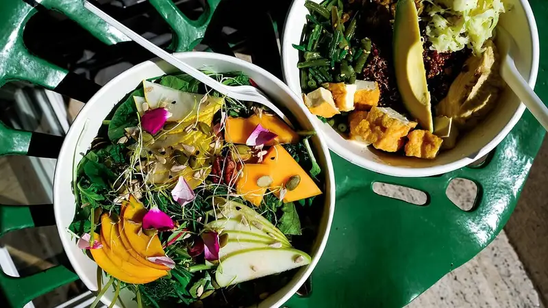 Dua mangkuk salad berisi sayuran hijau, irisan buah berwarna oranye, pir, alpukat, buncis, quinoa, dan potongan tahu di atas meja hijau.