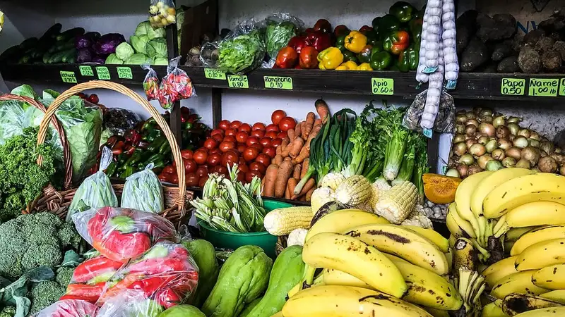 Lapak pasar berisi pisang, brokoli, tomat, wortel, jagung, dan sayuran hijau segar.