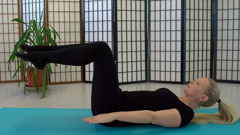 Seorang perempuan berbaring di matras biru melakukan gerakan pilates the hundred dengan kedua kaki terangkat dan ditekuk