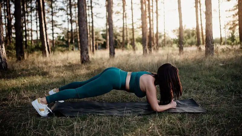 Seorang perempuan berpakaian olahraga hijau tosca menahan posisi plank bertumpu lengan bawah di atas matras di hutan pinus