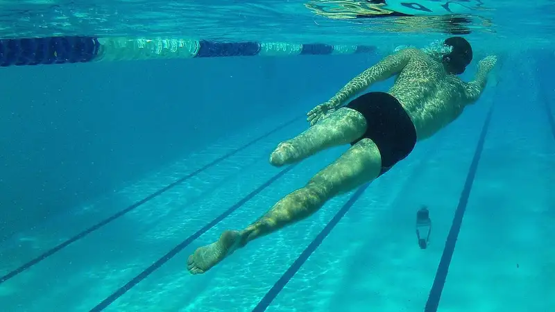 Seorang pria dewasa berenang gaya bebas di bawah permukaan air kolam renang, terlihat dari bawah air dengan garis lintasan di dasar kolam