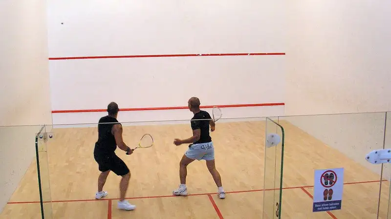 Dua pria bermain squash di lapangan dalam ruangan berlantai kayu dengan dinding putih bergaris merah.