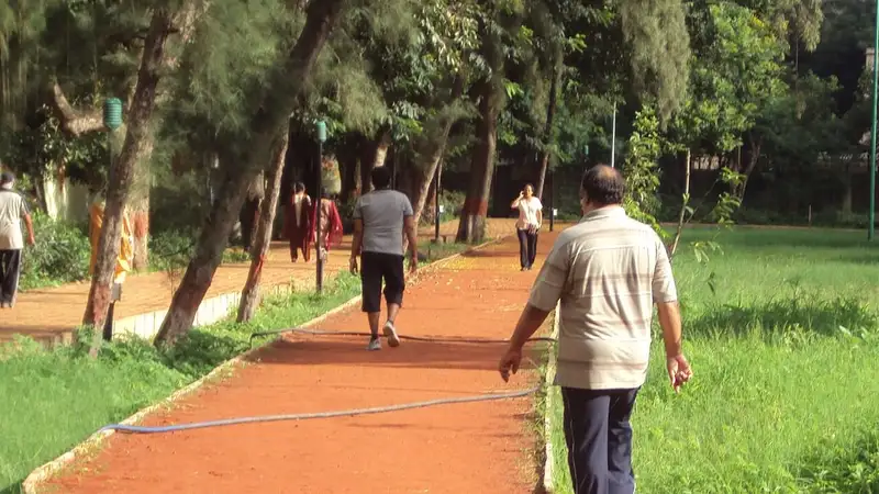 Beberapa orang berjalan kaki di jalur lari berwarna merah bata di sebuah taman berpohon di Pune, India, sebagian besar terlihat dari belakang.