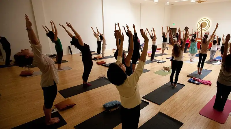 Sekelompok peserta kelas yoga berdiri di atas matras sambil mengangkat kedua tangan dan melengkungkan punggung ke belakang.