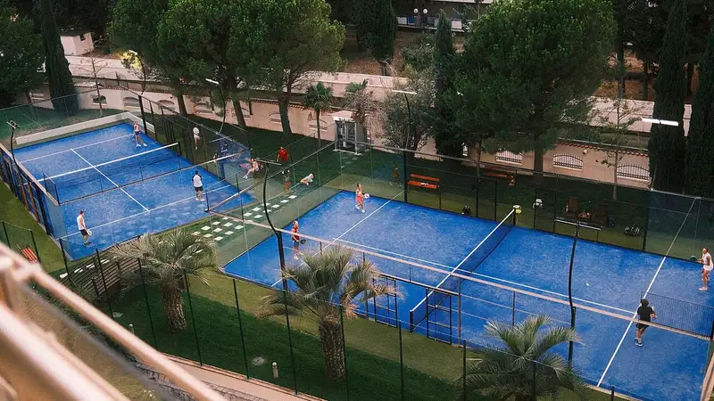 Beberapa lapangan padel biru berdinding kaca di area terbuka yang dikelilingi pepohonan, dengan pemain sedang bertanding