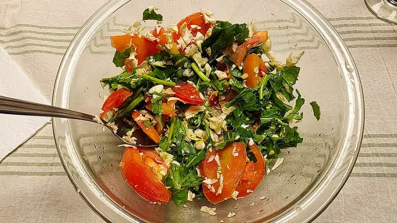 Salad segar berisi daun arugula, kol, dan potongan tomat di dalam mangkuk kaca bening dengan sendok saji.