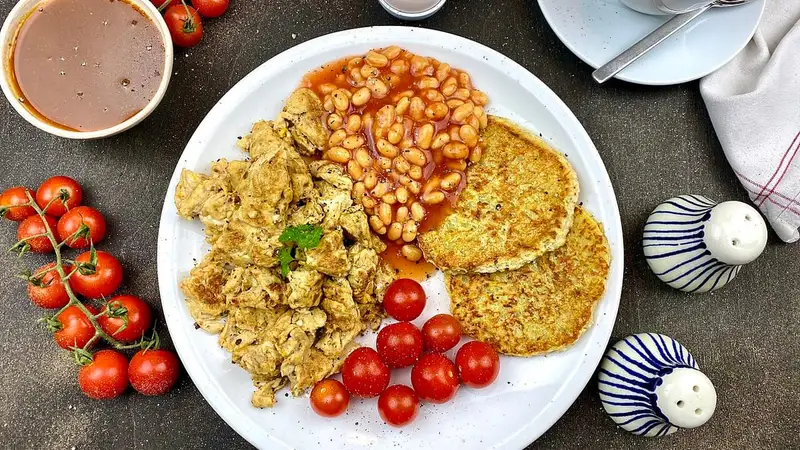 Piring sarapan berisi orak-arik, kacang panggang saus tomat, dua lembar rösti kentang, dan tomat ceri segar di atas meja gelap.