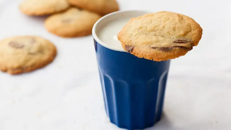 Segelas susu dalam gelas biru dengan kue kering cokelat di bibir gelas dan beberapa kue lain di atas meja putih
