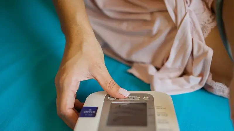 Tangan perempuan menekan tombol alat pengukur tekanan darah digital yang diletakkan di atas kasur.