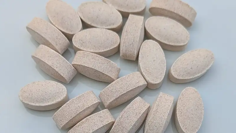 Tumpukan tablet suplemen vitamin C berbentuk oval berwarna krem bertekstur bintik di atas permukaan abu-abu terang.