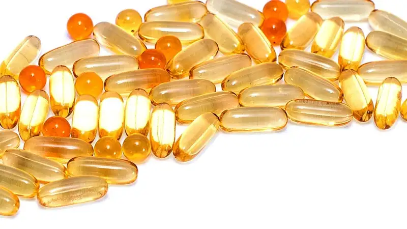 Tumpukan kapsul lunak berisi minyak berwarna kuning dan oranye di atas permukaan putih, bentuk suplemen yang umum untuk vitamin D.