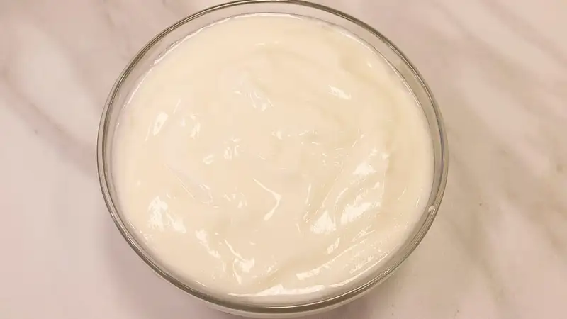 Mangkuk kaca bening berisi yogurt tawar putih yang kental, difoto dari atas di permukaan marmer.