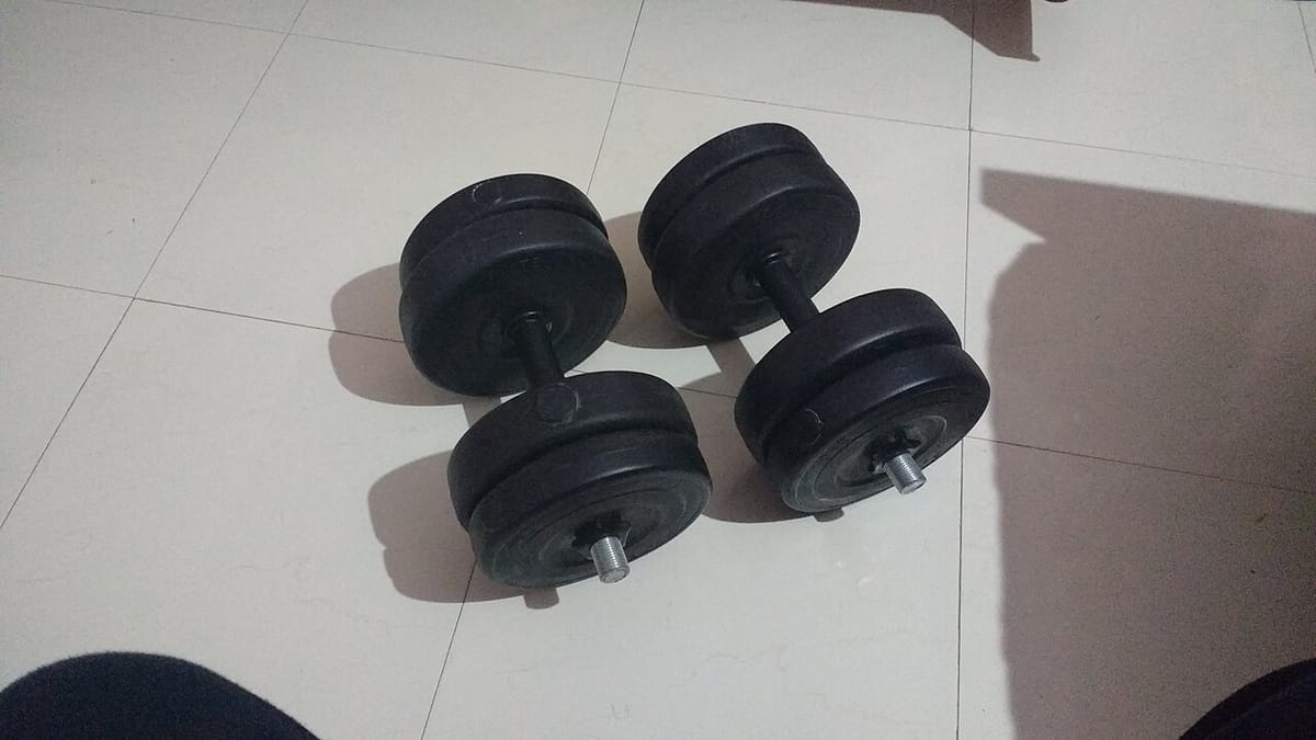 Sepasang dumbel hitam dengan piringan beban yang bisa dilepas pasang tergeletak di lantai keramik putih.