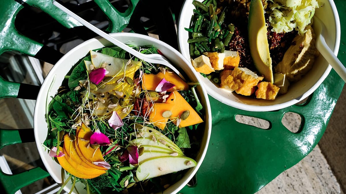 Dua mangkuk salad berisi sayuran hijau, irisan buah berwarna oranye, pir, alpukat, buncis, quinoa, dan potongan tahu di atas meja hijau.