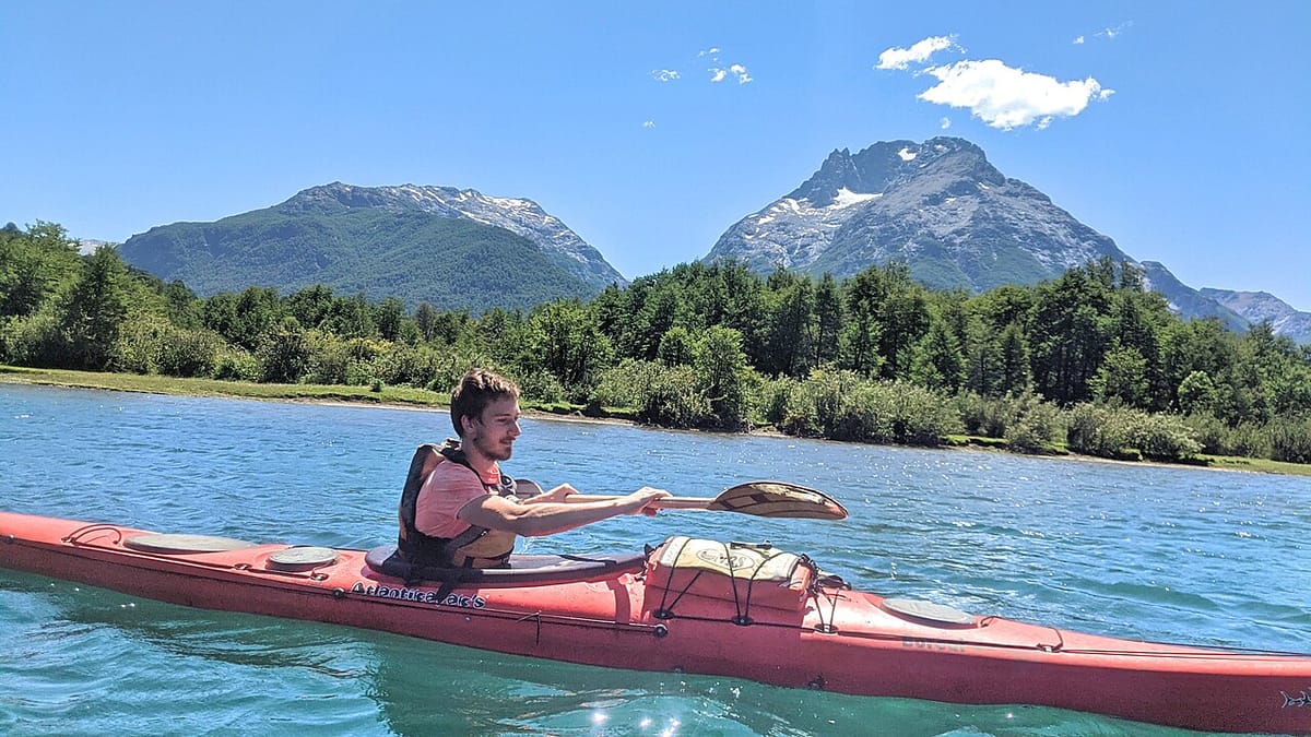 Seorang pria mendayung kayak touring merah di danau biru jernih dengan latar hutan dan pegunungan berpuncak salju