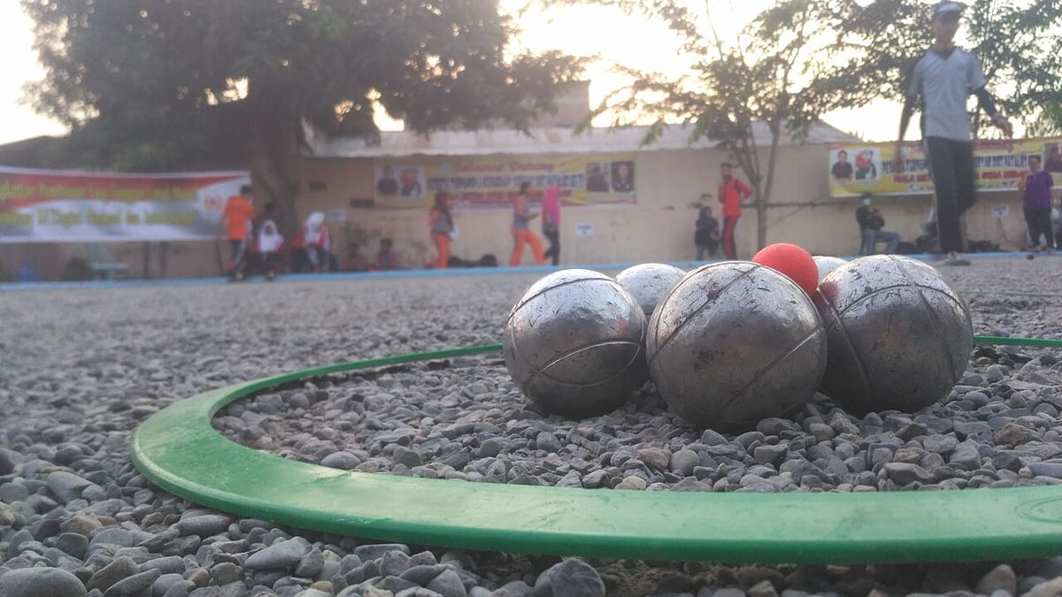 Beberapa bola besi petanque dan bola sasaran merah di dalam lingkaran lempar hijau di lapangan kerikil saat kejuaraan pelajar.