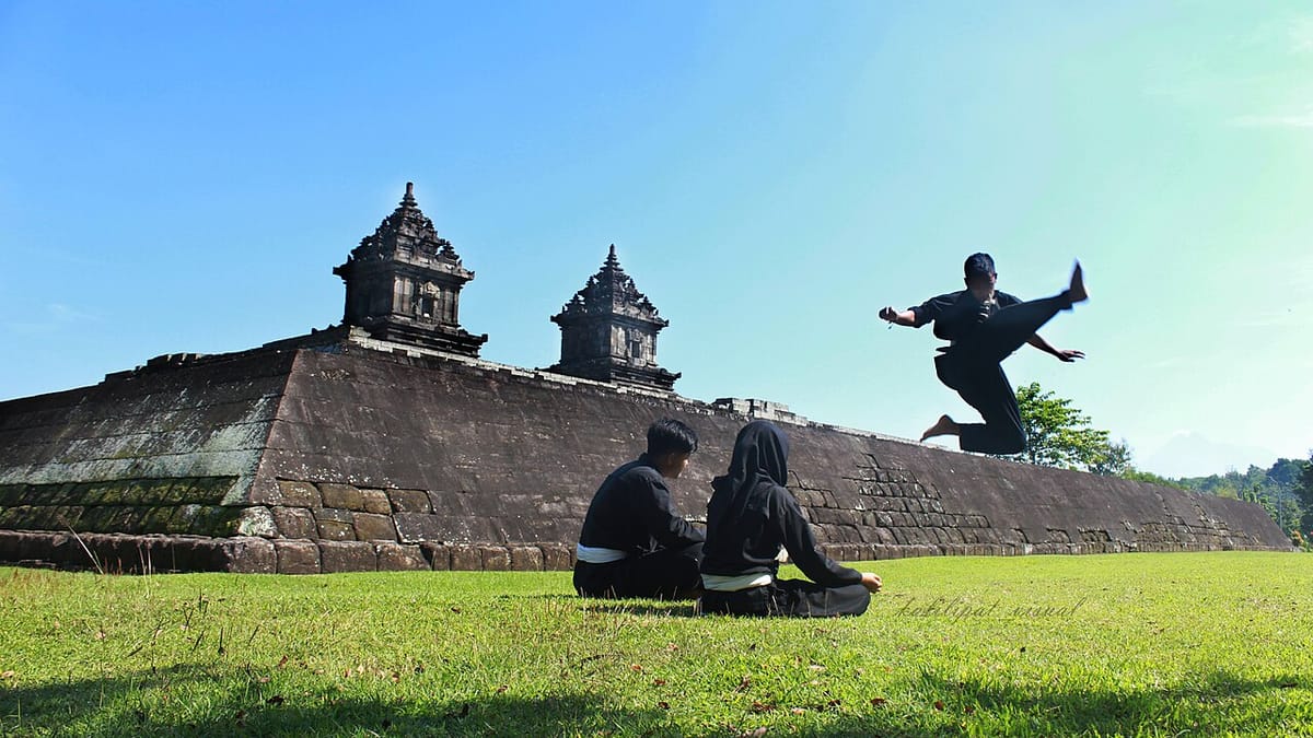 Pesilat berseragam hitam melakukan tendangan terbang di halaman rumput Candi Barong, Sleman, disaksikan dua rekannya