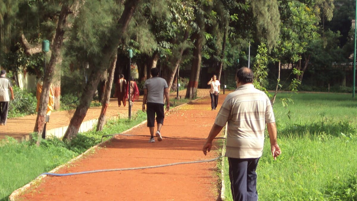 Beberapa orang berjalan kaki di jalur lari berwarna merah bata di sebuah taman berpohon di Pune, India, sebagian besar terlihat dari belakang.