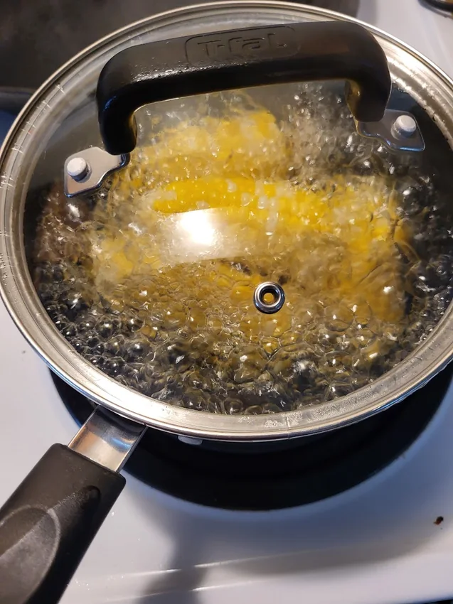Potongan tongkol jagung kuning direbus dalam panci kecil bertutup kaca di atas kompor.
