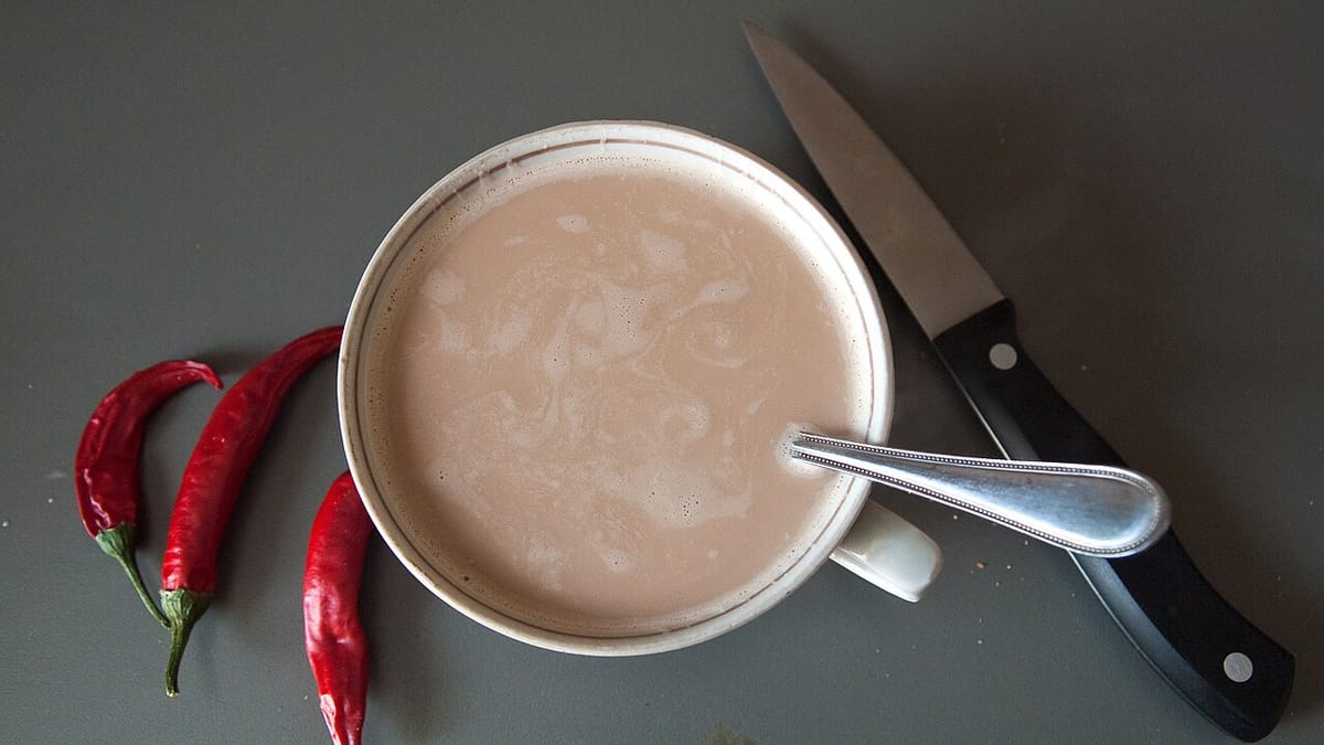 Secangkir kopi susu dengan sendok dan pisau di sampingnya, ditemani tiga cabai merah di atas meja abu-abu.