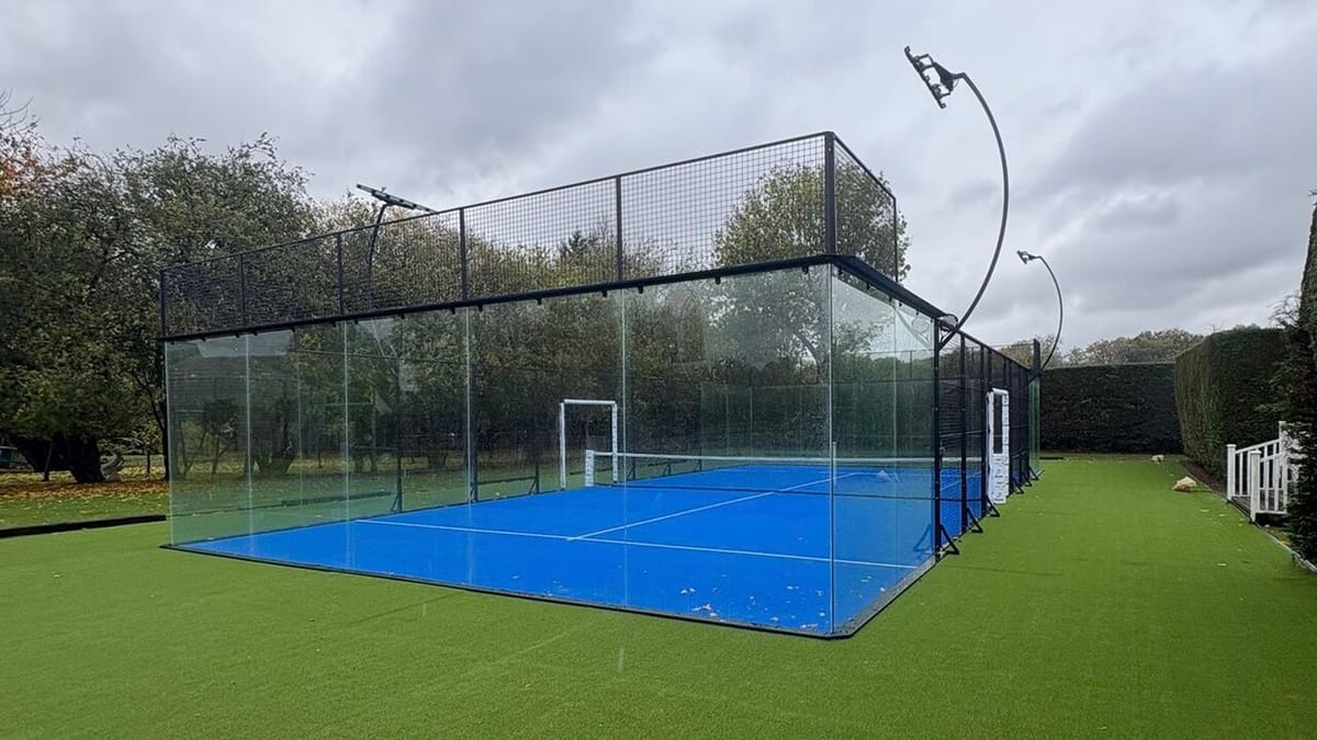 Lapangan padel outdoor berlantai biru yang dikelilingi dinding kaca dan pagar kawat, dengan net di tengah lapangan
