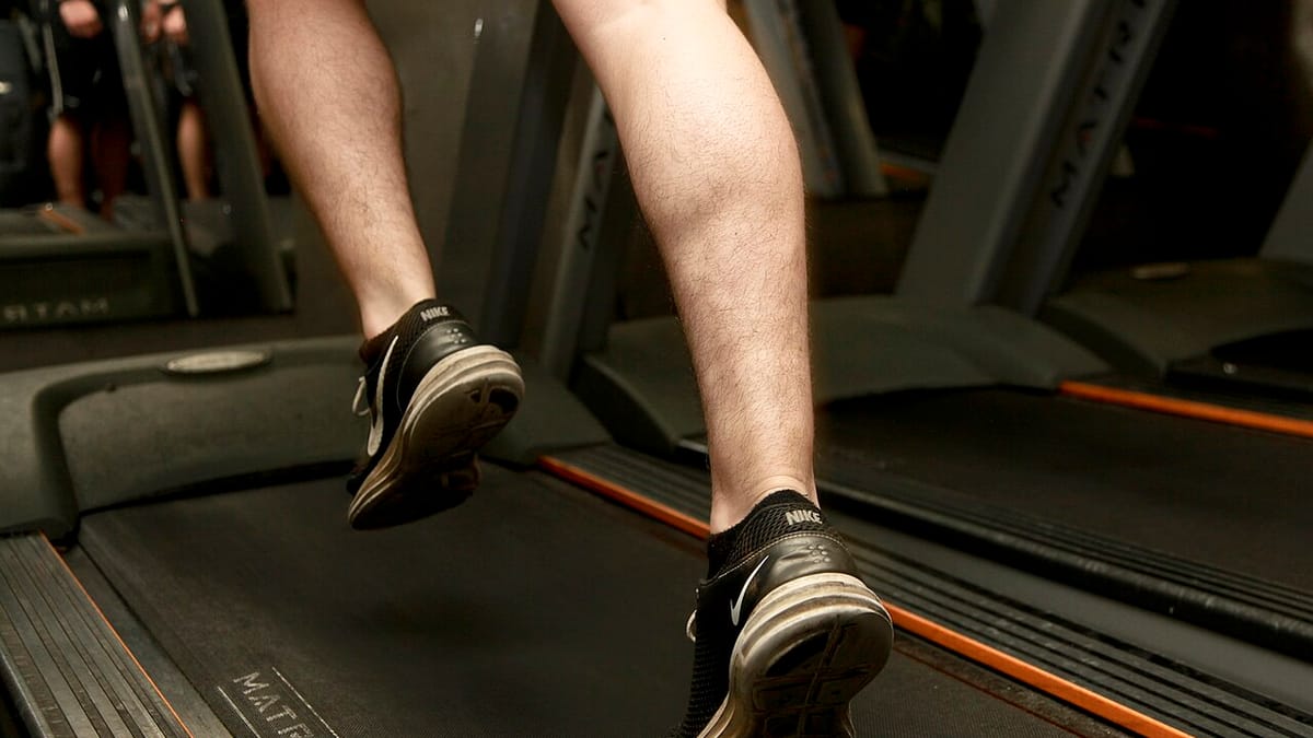 Kaki seorang pria bersepatu lari hitam sedang berlari di atas treadmill di ruang gym.
