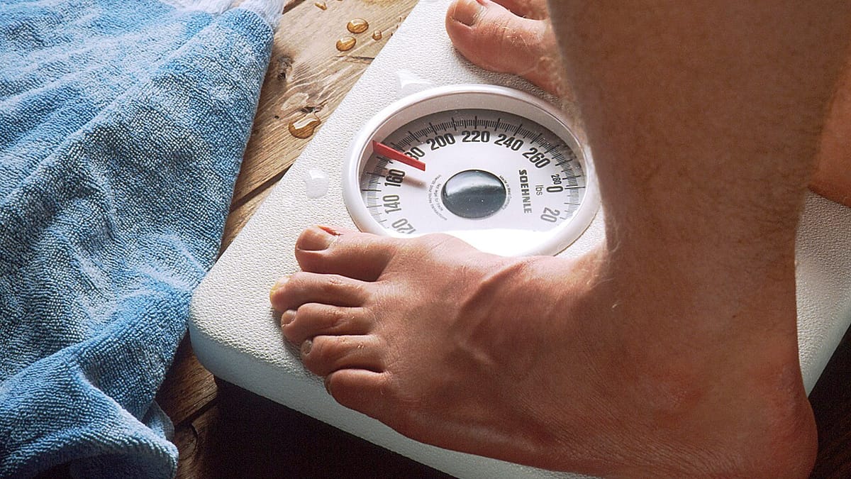 Kaki seseorang berdiri di atas timbangan badan analog di samping handuk biru di lantai kamar mandi.