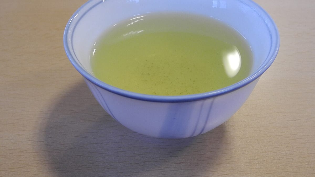 Mangkuk teh keramik putih bergaris biru berisi teh hijau berwarna kuning kehijauan di atas meja.