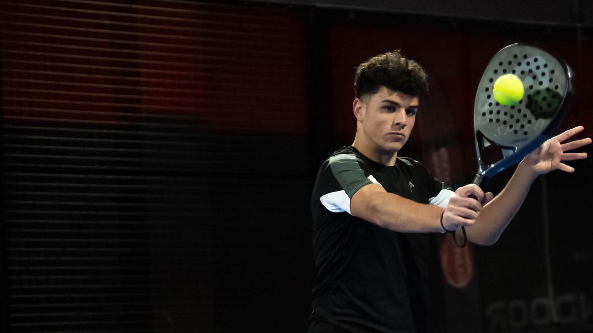 Pemain padel berkaus hitam memukul voli backhand di depan badan, bola kuning tepat di depan muka raket, dengan latar belakang gelap di dalam ruangan
