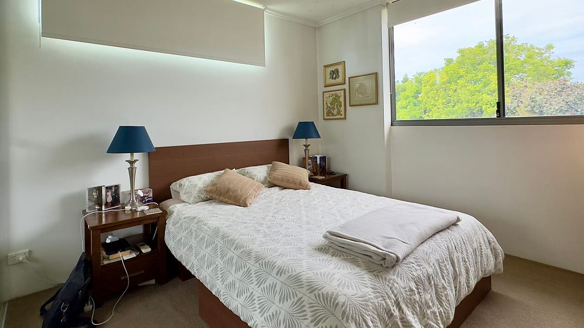 Kamar tidur yang rapi dengan ranjang berseprai putih bermotif daun, bantal-bantal, lampu meja biru di kedua sisi ranjang, dan cahaya siang dari jendela besar.